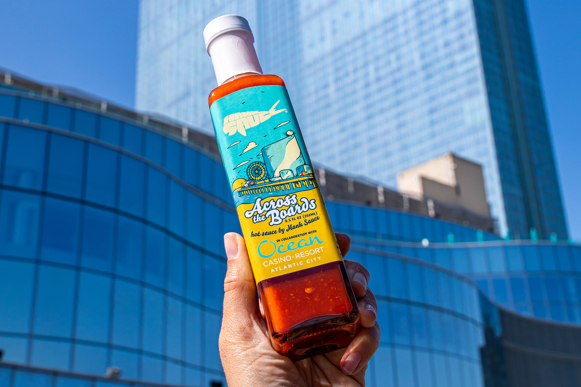 Hand holding a bottle of “Across the Boards” hot sauce, a Hank Sauce and Ocean Casino Resort collaboration, with the resort building in background.