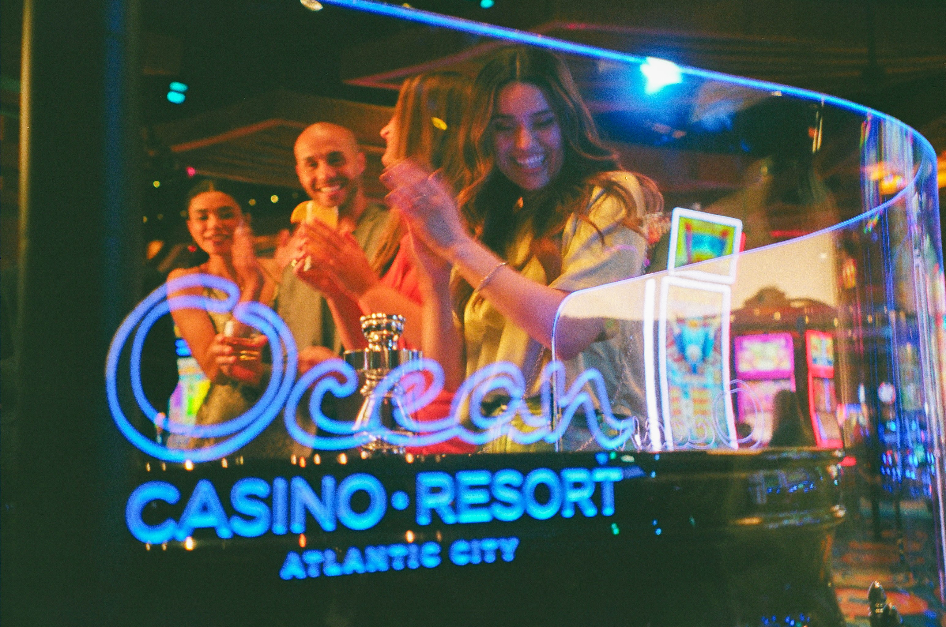 A group of friends claps and smiles behind a roulette table at Ocean Casino Resort, seen through glowing blue neon signage.