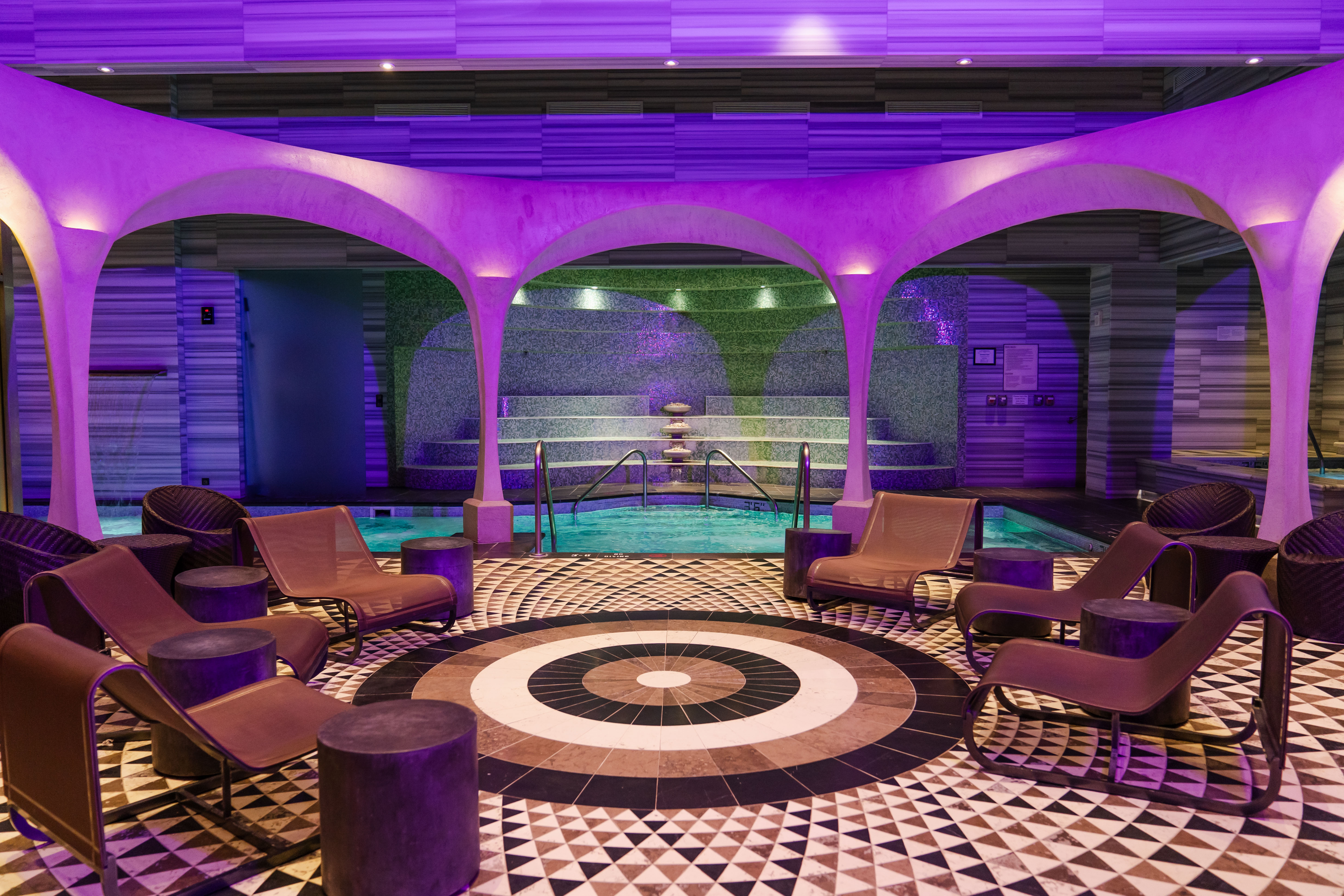 Indoor spa bathhouse with heated pool, lounge chairs, and arched architecture glowing with soft purple lighting